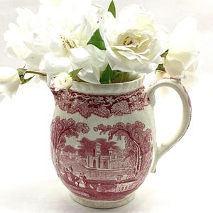May include: A ceramic pitcher featuring a deep red transferware pattern of a landscape with figures and a building, accented by a floral border. The pitcher is filled with a bouquet of white roses, showcasing a curved handle and a scalloped rim.