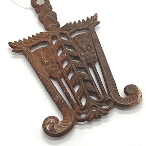 May include: Ornate, cast iron trivet in a dark brown color. The trivet features a decorative design with two brush-like shapes, leafy vines, and scrollwork. It has a handle with a hole for hanging.