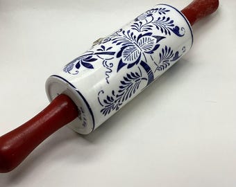 May include: A white ceramic rolling pin with a blue floral design and red wooden handles. The rolling pin is designed for baking and features a classic shape and decorative pattern.