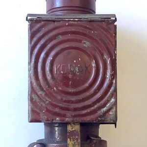 May include: A vintage maroon metal lantern featuring a square body with concentric circular patterns and the embossed text KOROK on the front. The lantern has a cylindrical top section, a flared base, and a small side handle, showing signs of wear.