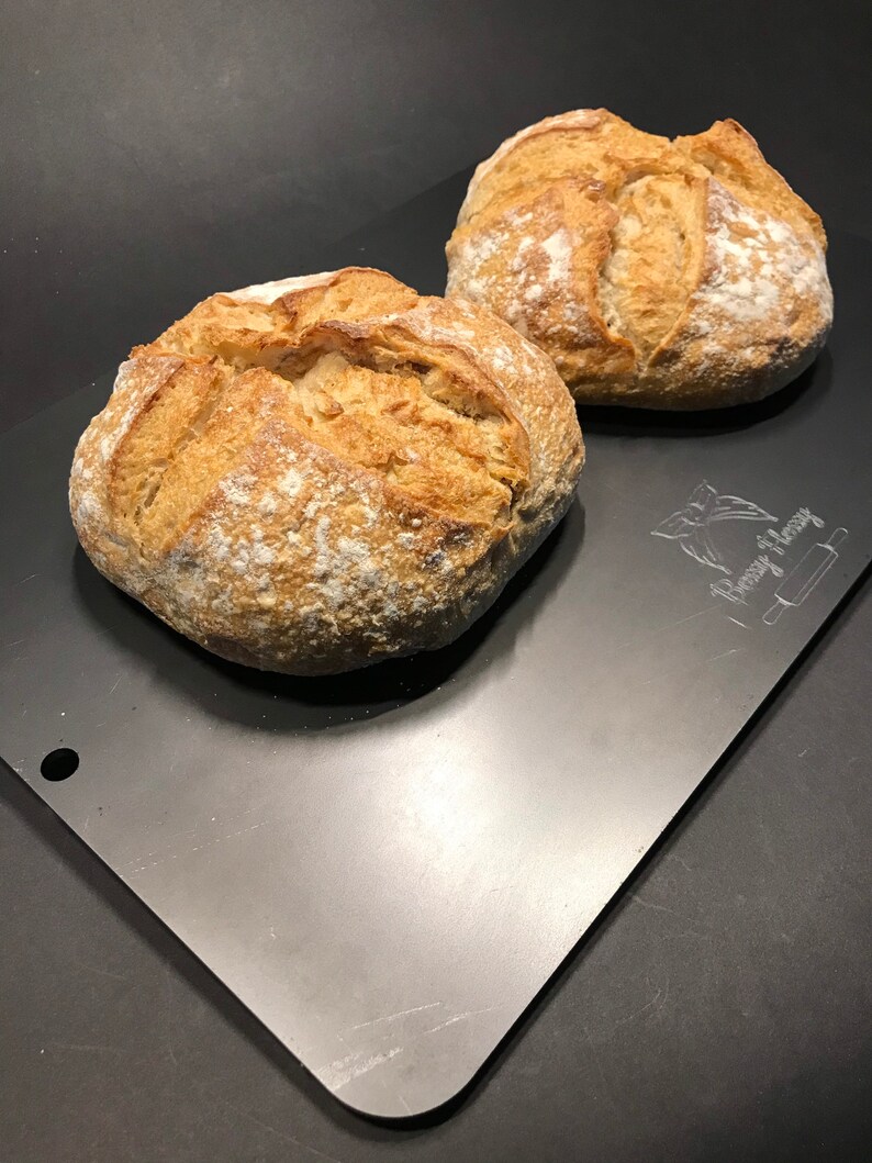 K&ouml;nnte beinhalten: Zwei frisch gebackene Sauerteigbrote auf einem silbernen Backblech. Das Brot ist goldbraun und hat eine leicht knusprige Kruste.