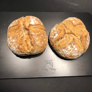 K&ouml;nnte beinhalten: Zwei runde Brote, die frisch gebacken auf einem schwarzen Backblech liegen. Das Brot hat eine goldbraune Kruste und eine leicht raue Textur.