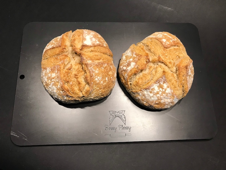 K&ouml;nnte beinhalten: Zwei runde Brote, die frisch gebacken auf einem schwarzen Backblech liegen. Das Brot hat eine goldbraune Kruste und eine leicht raue Textur.