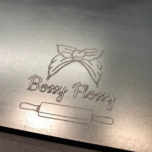 K&ouml;nnte beinhalten: Ein silbernes Metallschild mit einer Schwarzwei&szlig;illustration einer Haarschleife und dem Text "Bossy Flossy". Unter dem Text befindet sich eine Schwarzwei&szlig;illustration eines Nudelholzes.