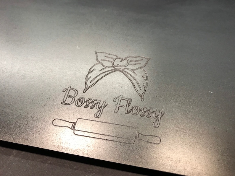 K&ouml;nnte beinhalten: Ein silbernes Metallschild mit einer Schwarzwei&szlig;illustration einer Haarschleife und dem Text "Bossy Flossy". Unter dem Text befindet sich eine Schwarzwei&szlig;illustration eines Nudelholzes.