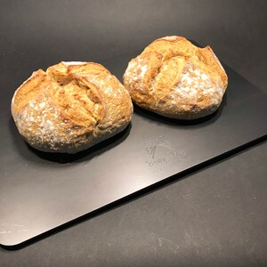 K&ouml;nnte beinhalten: Zwei runde Brote, frisch gebacken, auf einem schwarzen Backblech aus Metall. Das Brot ist leicht mit Mehl best&auml;ubt und hat eine goldbraune Kruste.