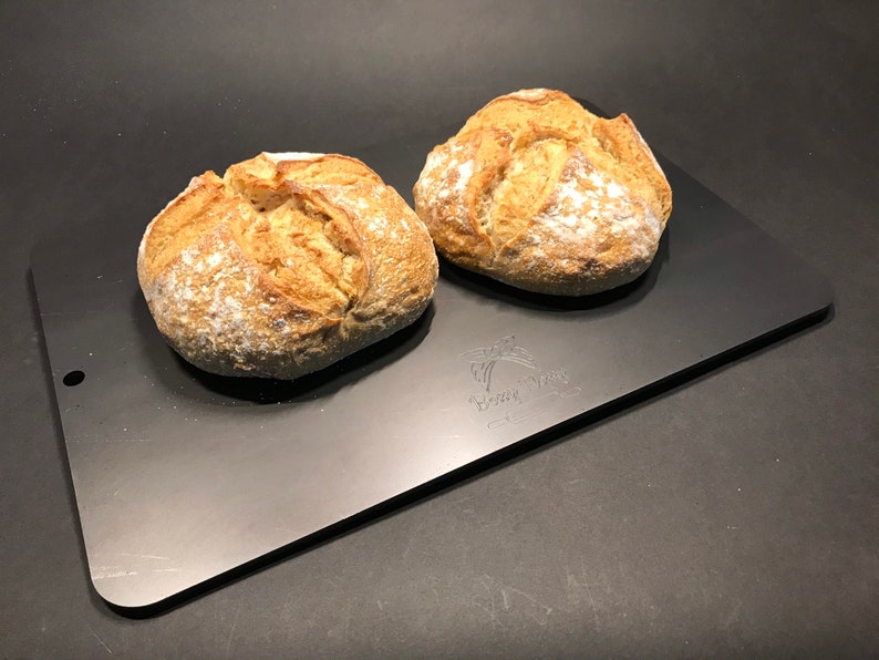 K&ouml;nnte beinhalten: Zwei runde Brote, frisch gebacken, auf einem schwarzen Backblech aus Metall. Das Brot ist leicht mit Mehl best&auml;ubt und hat eine goldbraune Kruste.