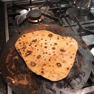 K&ouml;nnte beinhalten: Eine runde, schwarze Gusseisenpfanne mit einem braunen, flachen Brot darauf gebacken. Die Pfanne hat einen erh&ouml;hten Rand und eine strukturierte Oberfl&auml;che.