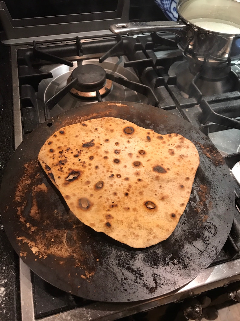 K&ouml;nnte beinhalten: Eine runde, schwarze Gusseisenpfanne mit einem braunen, flachen Brot darauf gebacken. Die Pfanne hat einen erh&ouml;hten Rand und eine strukturierte Oberfl&auml;che.