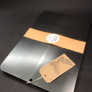 K&ouml;nnte beinhalten: Ein gro&szlig;es, rechteckiges, silbernes Backblech aus Metall mit abgerundeten Ecken. Das Blech ist in braunes Papier eingewickelt, mit einem wei&szlig;en runden Aufkleber, auf dem "Betty Flamingo" steht. Ein braunes Papieretikett mit einem Schmetterlingsdesign ist am Blech befestigt.