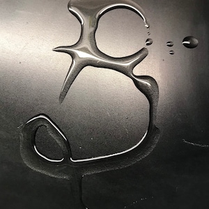 K&ouml;nnte beinhalten: Nahaufnahme einer schwarzen Oberfl&auml;che mit einem Wassertropfen, der die Form des Buchstabens "B" gebildet hat.