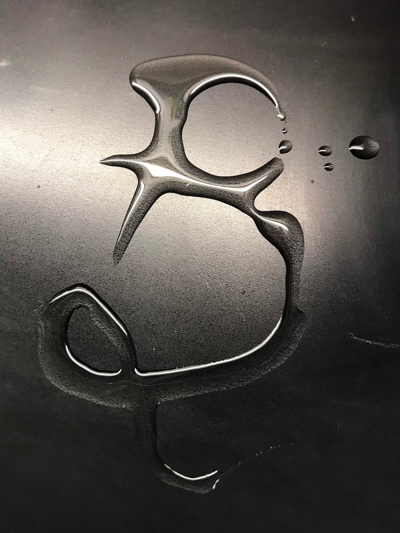 K&ouml;nnte beinhalten: Nahaufnahme einer schwarzen Oberfl&auml;che mit einem Wassertropfen, der die Form des Buchstabens "B" gebildet hat.