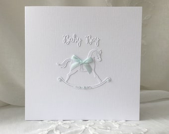 May include: A white square greeting card with the words "Baby Boy" in script. A white embossed rocking horse with a light blue bow is centered below the text. The card has a textured surface.