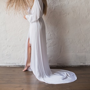 Op de afbeelding: Een vrouw in een witte maxi-jurk met een lange sleep staat voor een witte muur. Ze houdt een grote, ronde, geweven wanddecoratie vast.