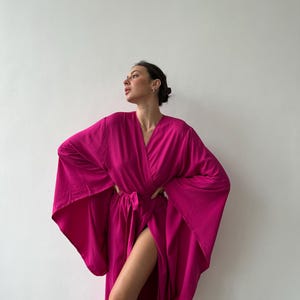 May include: A vibrant fuchsia-colored robe with wide sleeves and a wrap-around design. The robe is tied at the waist with a matching belt. The fabric appears lightweight and flowing, suggesting a comfortable garment. The robe is worn open, revealing a high slit.