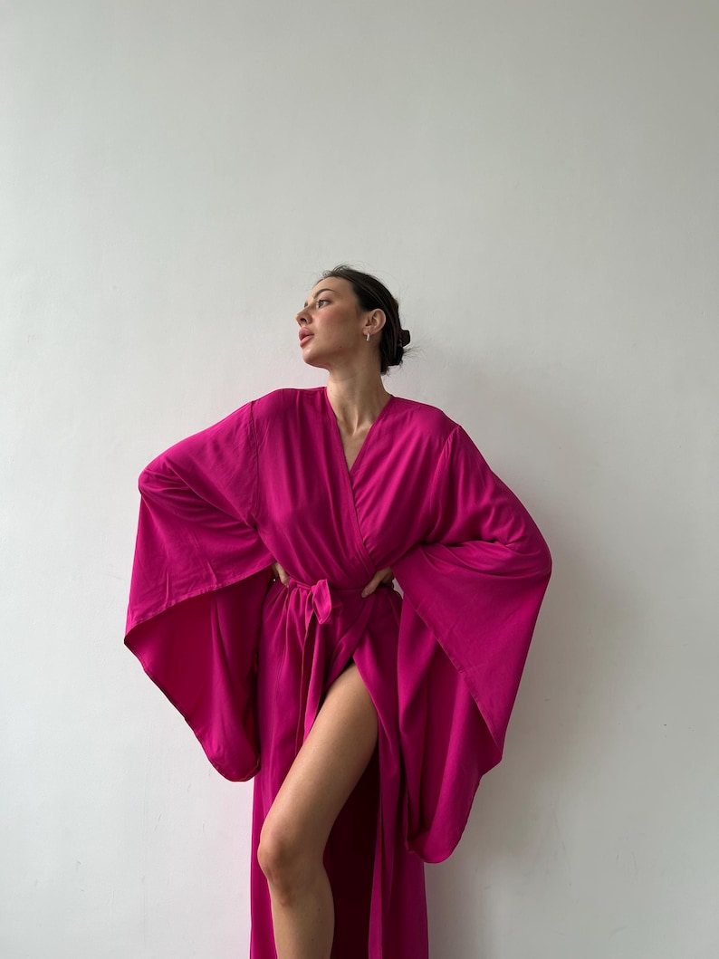 May include: A vibrant fuchsia-colored robe with wide sleeves and a wrap-around design. The robe is tied at the waist with a matching belt. The fabric appears lightweight and flowing, suggesting a comfortable garment. The robe is worn open, revealing a high slit.
