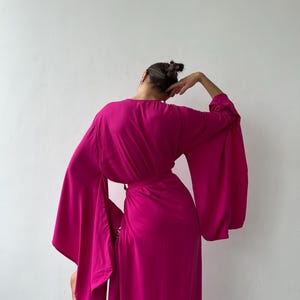 May include: A magenta-colored wrap dress with wide, flowing sleeves. The dress is cinched at the waist with a matching belt. The model is turned away from the camera, with their arm raised. The dress is made of a soft, draping fabric.