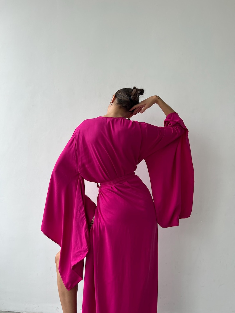 May include: A magenta-colored wrap dress with wide, flowing sleeves. The dress is cinched at the waist with a matching belt. The model is turned away from the camera, with their arm raised. The dress is made of a soft, draping fabric.