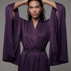 Peut inclure: Une robe en satin violet foncé avec de larges manches et une ceinture assortie nouée à la taille. La robe a un col en V et est faite d'un tissu lisse et fluide. Le modèle porte la robe et a les mains sur la tête.