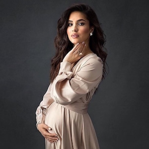 May include: A pregnant woman wearing a long, light beige satin dress with long sleeves. She is standing with her hand on her stomach and looking at the camera.