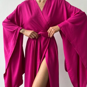May include: A vibrant fuchsia wrap dress with a V-neckline and wide, flowing sleeves. The dress features a high slit, showcasing a leg. The fabric appears soft and drapes elegantly, creating a sophisticated silhouette.