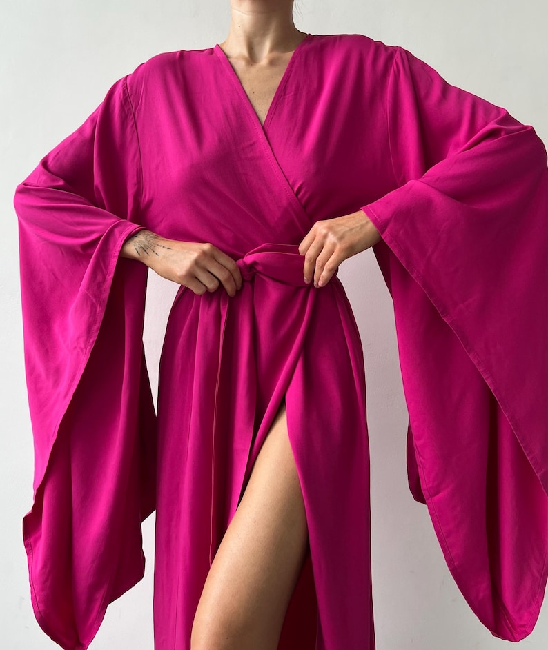 May include: A vibrant fuchsia wrap dress with a V-neckline and wide, flowing sleeves. The dress features a high slit, showcasing a leg. The fabric appears soft and drapes elegantly, creating a sophisticated silhouette.