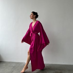 May include: A magenta-colored robe with wide sleeves and a wrap-around design. The robe is floor-length and features a belt at the waist. The fabric appears soft and flowing, creating a relaxed and elegant look. The robe is suitable for lounging or as a stylish cover-up.