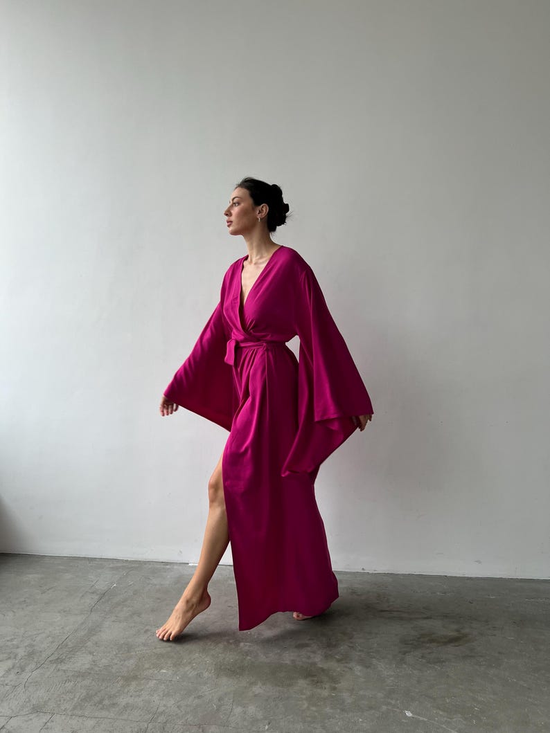 May include: A magenta-colored robe with wide sleeves and a wrap-around design. The robe is floor-length and features a belt at the waist. The fabric appears soft and flowing, creating a relaxed and elegant look. The robe is suitable for lounging or as a stylish cover-up.