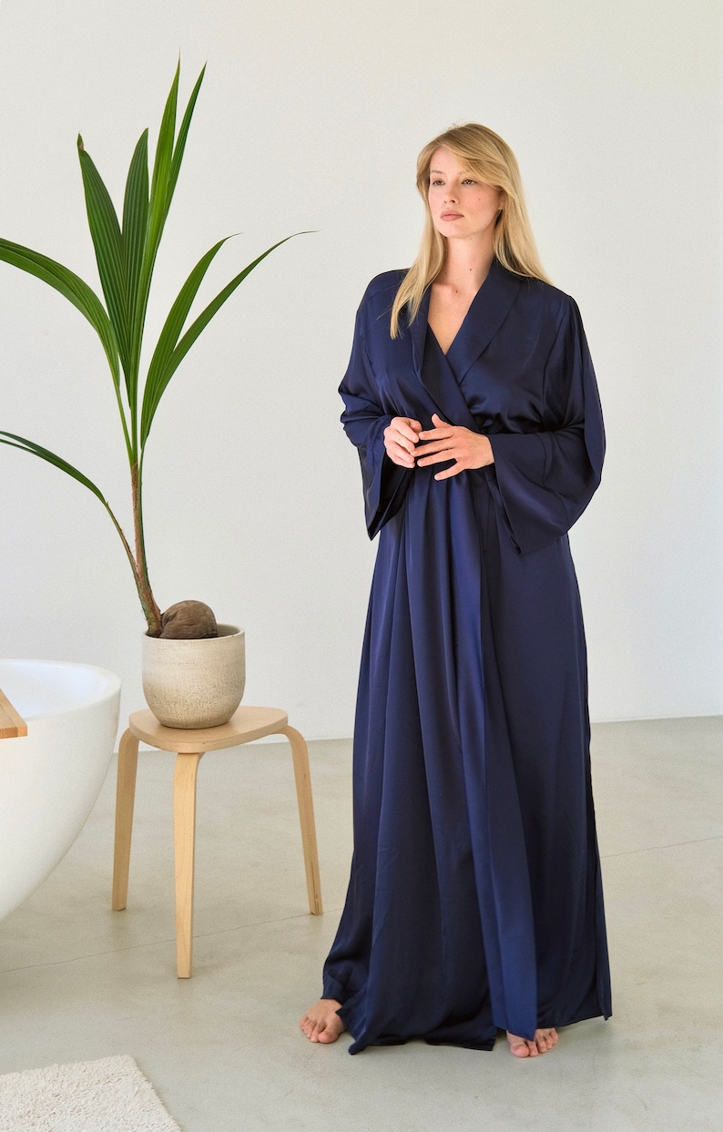 May include: A long, navy blue satin robe with a shawl collar and long sleeves. The robe is tied at the waist with a sash.