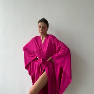 May include: A vibrant fuchsia-colored robe with wide sleeves and a wrap-around design. The robe is tied at the waist with a matching belt. The fabric appears soft and flowing, creating a dramatic silhouette. The robe is a solid color with a V-neckline.