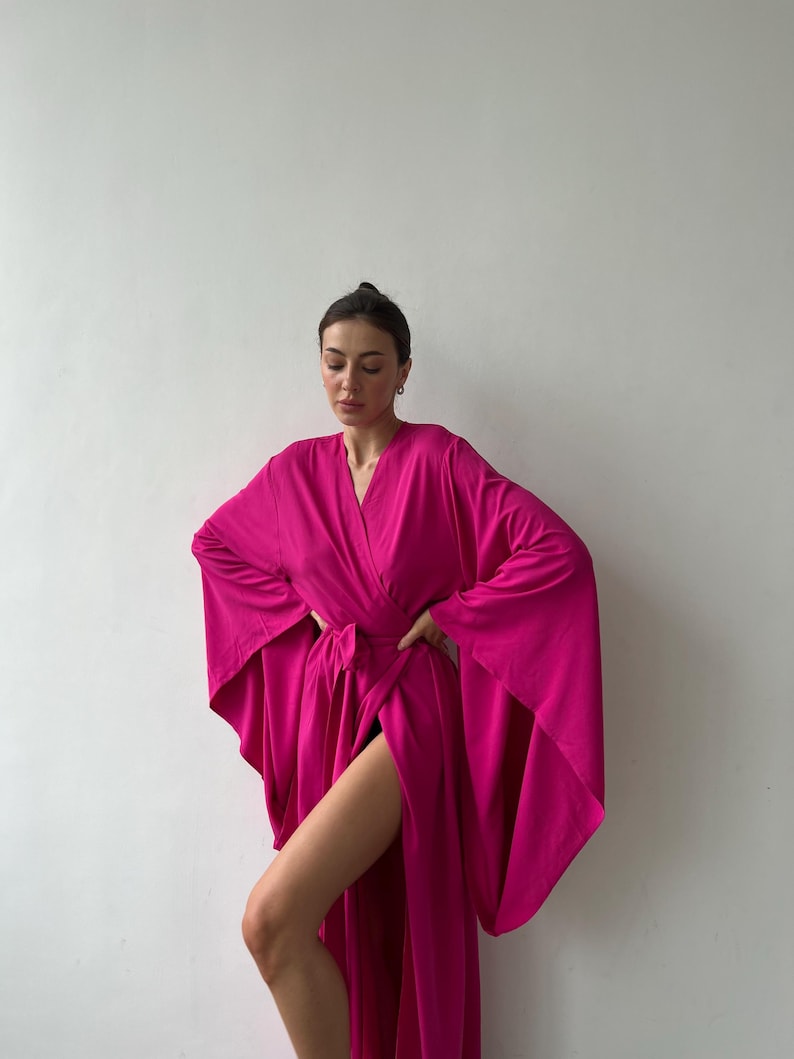 May include: A vibrant fuchsia-colored robe with wide sleeves and a wrap-around design. The robe is tied at the waist with a matching belt. The fabric appears soft and flowing, creating a dramatic silhouette. The robe is a solid color with a V-neckline.