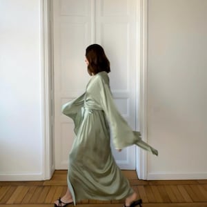 May include: A woman wearing a flowing sage green satin robe with long sleeves and a tie belt. She is walking in a room with wooden floors.