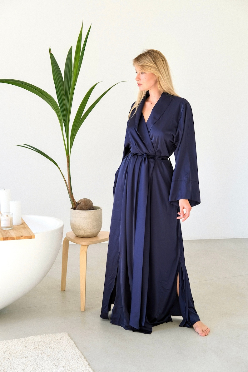 May include: A navy blue satin robe with long sleeves and a tie belt. The robe has side slits and a relaxed fit.