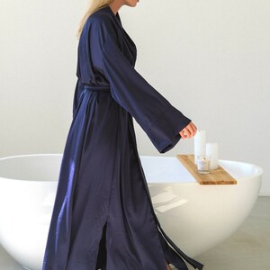 May include: A woman wearing a long, dark blue satin robe with a sash and a front slit. She is standing in a bathroom with a white bathtub and a wooden tray with candles on it.