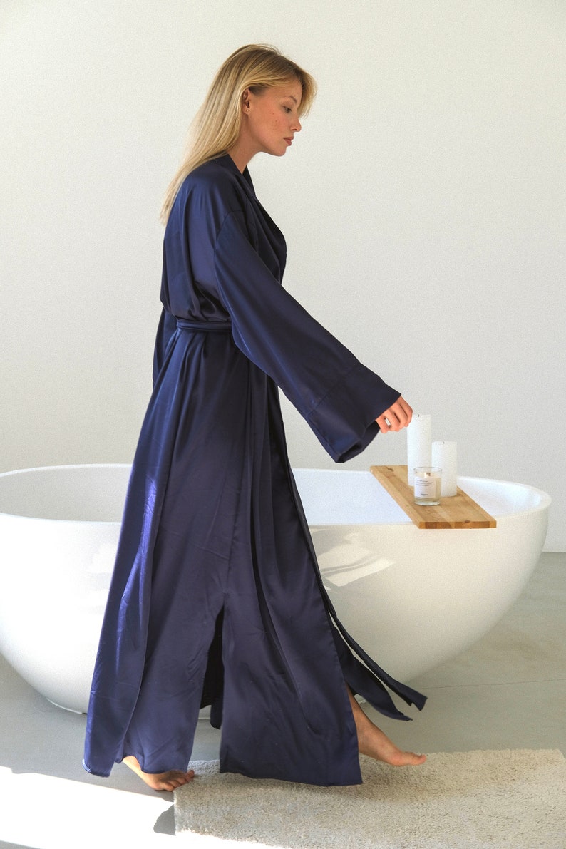May include: A woman wearing a long, dark blue satin robe with a sash and a front slit. She is standing in a bathroom with a white bathtub and a wooden tray with candles on it.
