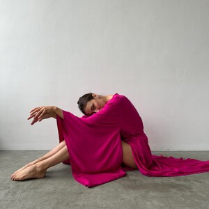 May include: A person wearing a flowing, magenta-colored robe with wide sleeves, seated on a gray surface. The robe drapes to the floor, creating a dramatic effect. The person is leaning forward, with their head resting on their arms.