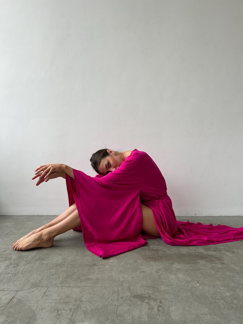 May include: A person wearing a flowing, magenta-colored robe with wide sleeves, seated on a gray surface. The robe drapes to the floor, creating a dramatic effect. The person is leaning forward, with their head resting on their arms.