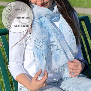 May include: A light blue scarf with a floral pattern and fringe detail. The scarf has UV protection, is antibacterial, and has a cool touch. The scarf is draped around the shoulders.