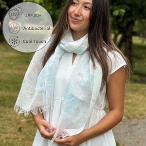 May include: A white and light blue scarf with fringe detail. The scarf has a light blue watercolor design and is draped around the neck. The image also includes a graphic with the text "UV UPF 30+", "Antibacterial", and "Cool Touch".