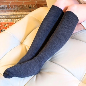 May include: A pair of dark gray knee-high socks. The socks are made of a textured knit fabric and are pulled up to the knees. The socks are on a cream-colored leather couch.
