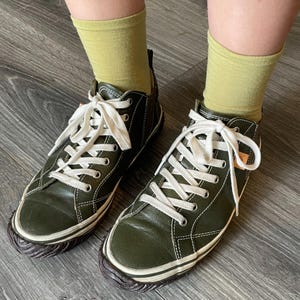 May include: A pair of olive green leather sneakers with white laces and white stitching details. The shoes feature a dark brown textured rubber toe cap and a matching dark brown rubber sole. The sneakers are worn with light green crew socks.