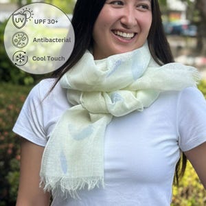 May include: A light yellow scarf with a subtle blue leaf pattern and fringed edges. The scarf features text indicating UPF 30+ protection, antibacterial properties, and a cool touch fabric finish, presented in a soft, lightweight material.