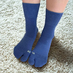 May include: A pair of blue tabi socks with a split toe design. The socks are a solid blue color with a ribbed cuff and the word "ILIR" printed on the side of each sock. The socks are worn on a cream-colored carpet.