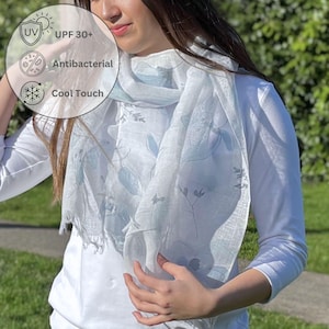 May include: A light blue and white scarf with a floral pattern is draped around a person's neck. The scarf has a fringed edge and is made of a lightweight material. The image also includes text that says "UV UPF 30+", "Antibacterial", and "Cool Touch."