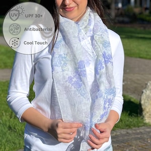 May include: A white long-sleeve shirt paired with a lightweight scarf featuring a blue and green floral pattern. The scarf includes text labels for UPF 30+, Antibacterial, and Cool Touch properties, indicating sun protection and cooling features.