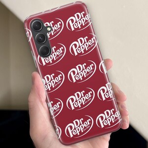 Puede incluir: Una funda de tel&eacute;fono burdeos con un patr&oacute;n repetido del logotipo blanco de Dr Pepper. La funda del tel&eacute;fono tiene una matriz de c&aacute;mara en la parte superior izquierda. El logotipo de Dr Pepper incluye las palabras "Est. 1885".