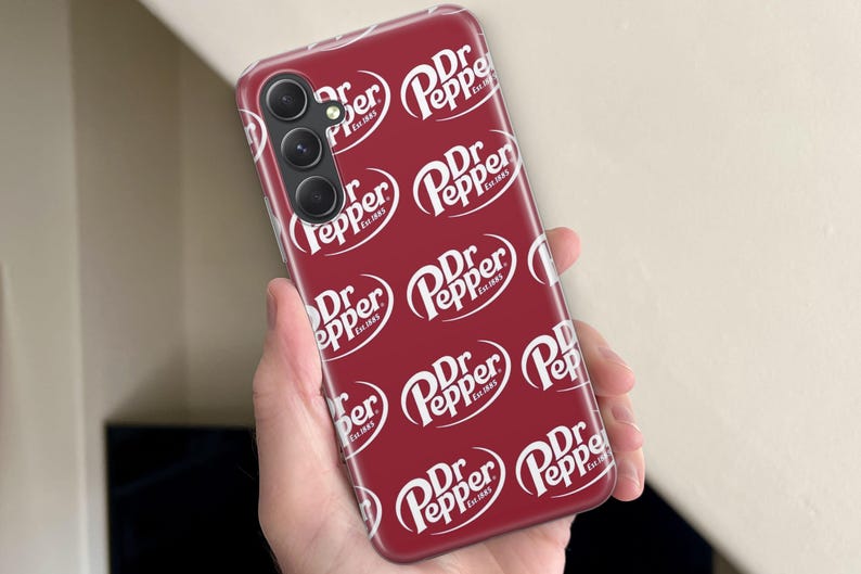 Puede incluir: Una funda de tel&eacute;fono burdeos con un patr&oacute;n repetido del logotipo blanco de Dr Pepper. La funda del tel&eacute;fono tiene una matriz de c&aacute;mara en la parte superior izquierda. El logotipo de Dr Pepper incluye las palabras "Est. 1885".