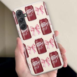 Puede incluir: Una funda de tel&eacute;fono con un patr&oacute;n repetido de latas de Dr. Pepper y lazos rosas sobre un fondo crema. Las latas de Dr. Pepper son de color rojo oscuro con texto blanco y el n&uacute;mero 23. Los lazos son de color rosa claro.