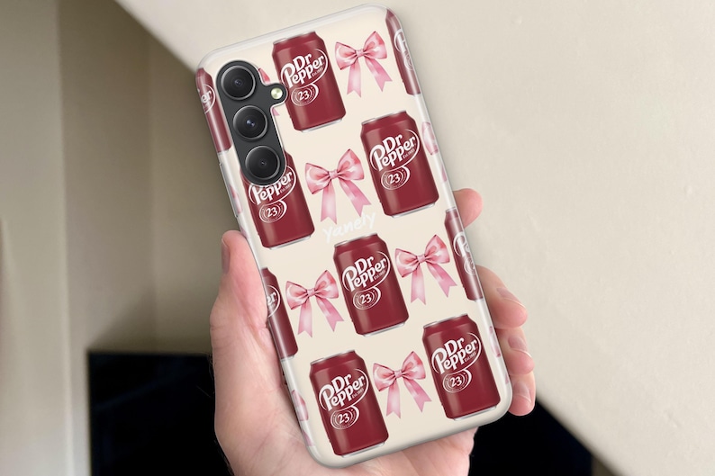 Puede incluir: Una funda de tel&eacute;fono con un patr&oacute;n repetido de latas de Dr. Pepper y lazos rosas sobre un fondo crema. Las latas de Dr. Pepper son de color rojo oscuro con texto blanco y el n&uacute;mero 23. Los lazos son de color rosa claro.