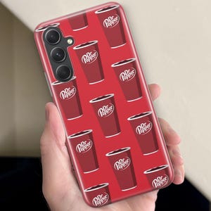 Puede incluir: Funda de tel&eacute;fono roja con un patr&oacute;n repetido de vasos de refresco Dr. Pepper. Los vasos son de color rojo oscuro con texto blanco y un borde blanco. La funda tiene una c&aacute;mara de triple lente.